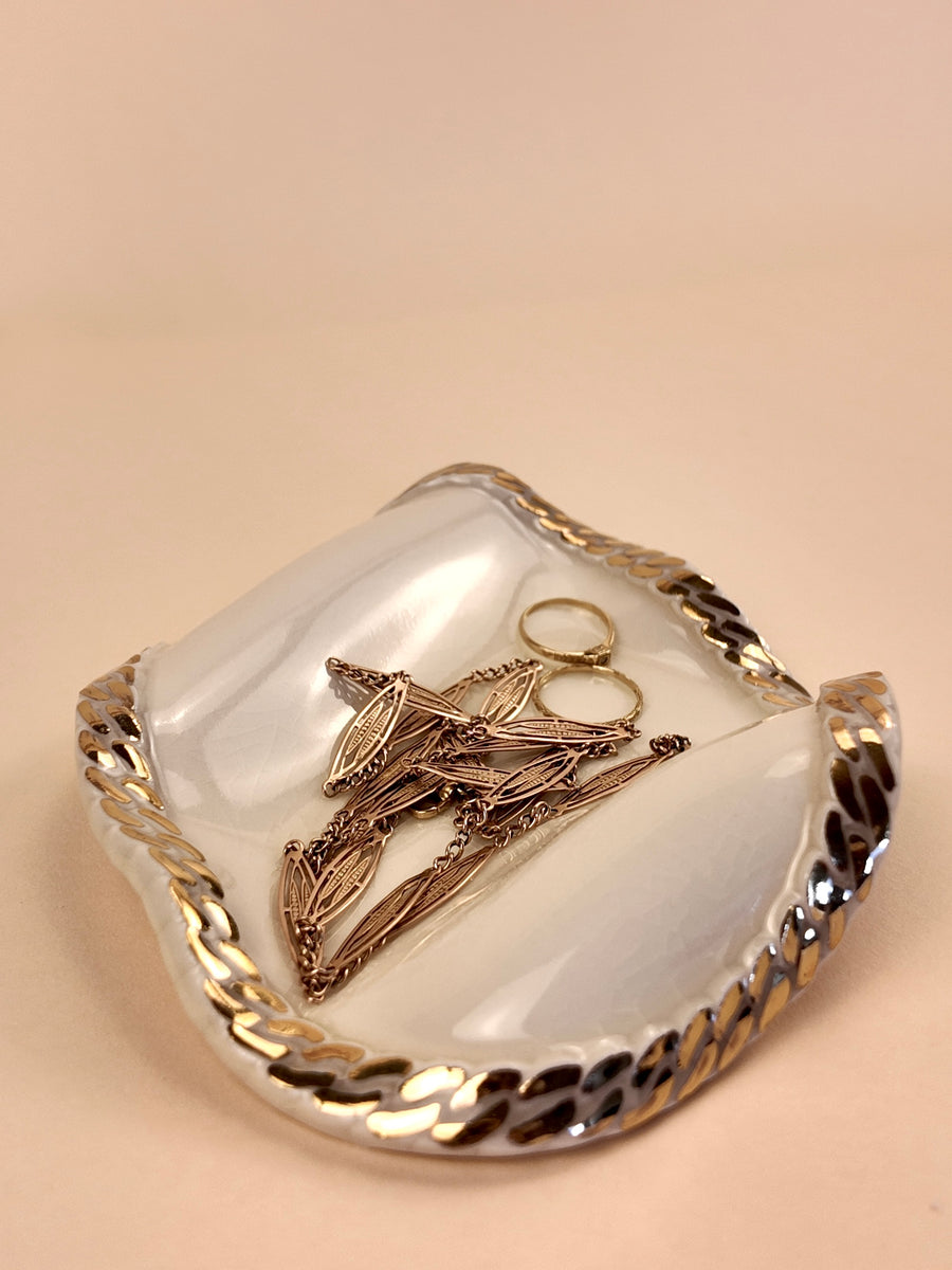 A white ceramic jewelry organizer tray with a braided gold and silver cuban chain trim, holding several gold-toned rings and earrings, set against a solid beige background.