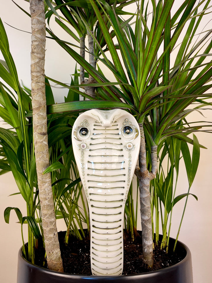 A porcelain garden statue shaped like a cobra snake head, displayed in a black planter among green potted plants.