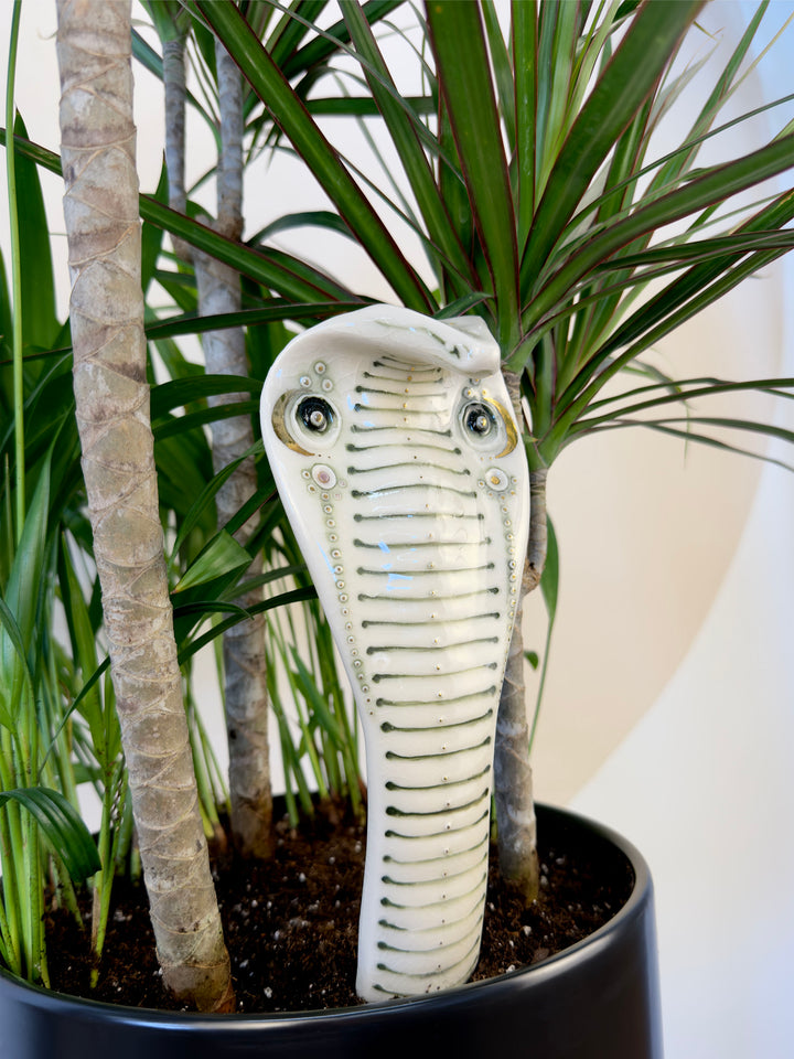 A porcelain garden statue shaped like a cobra snake head, displayed in a black planter among green potted plants.