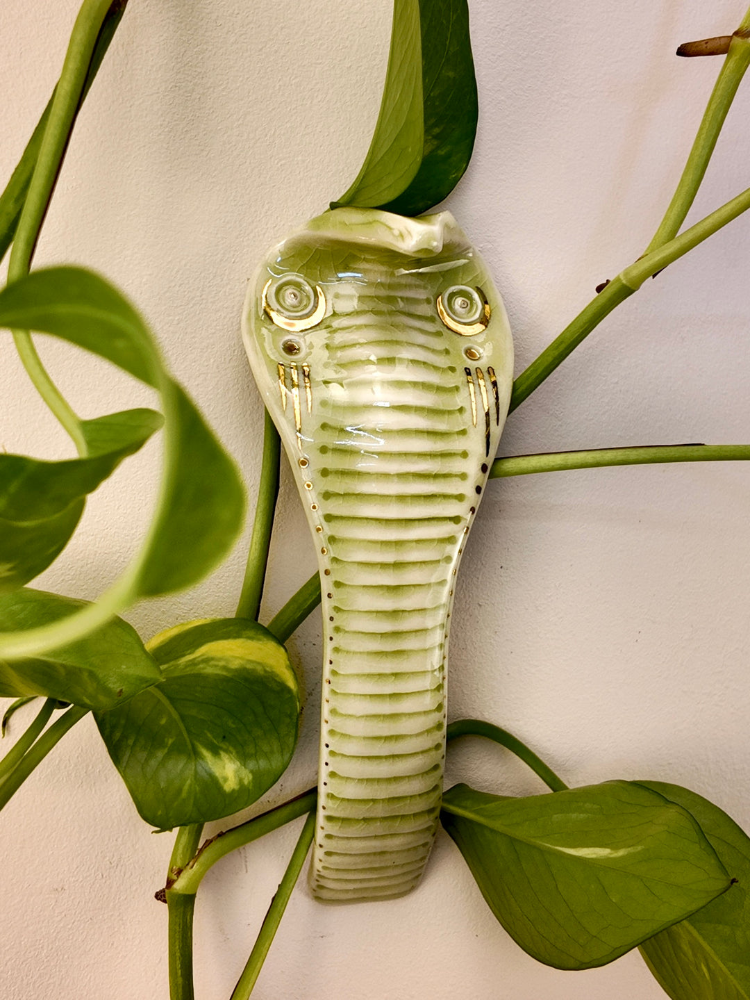 A ceramic wall-mounted snake figurine with a glossy finish, styled among green houseplant leaves against a plain off-white wall.
