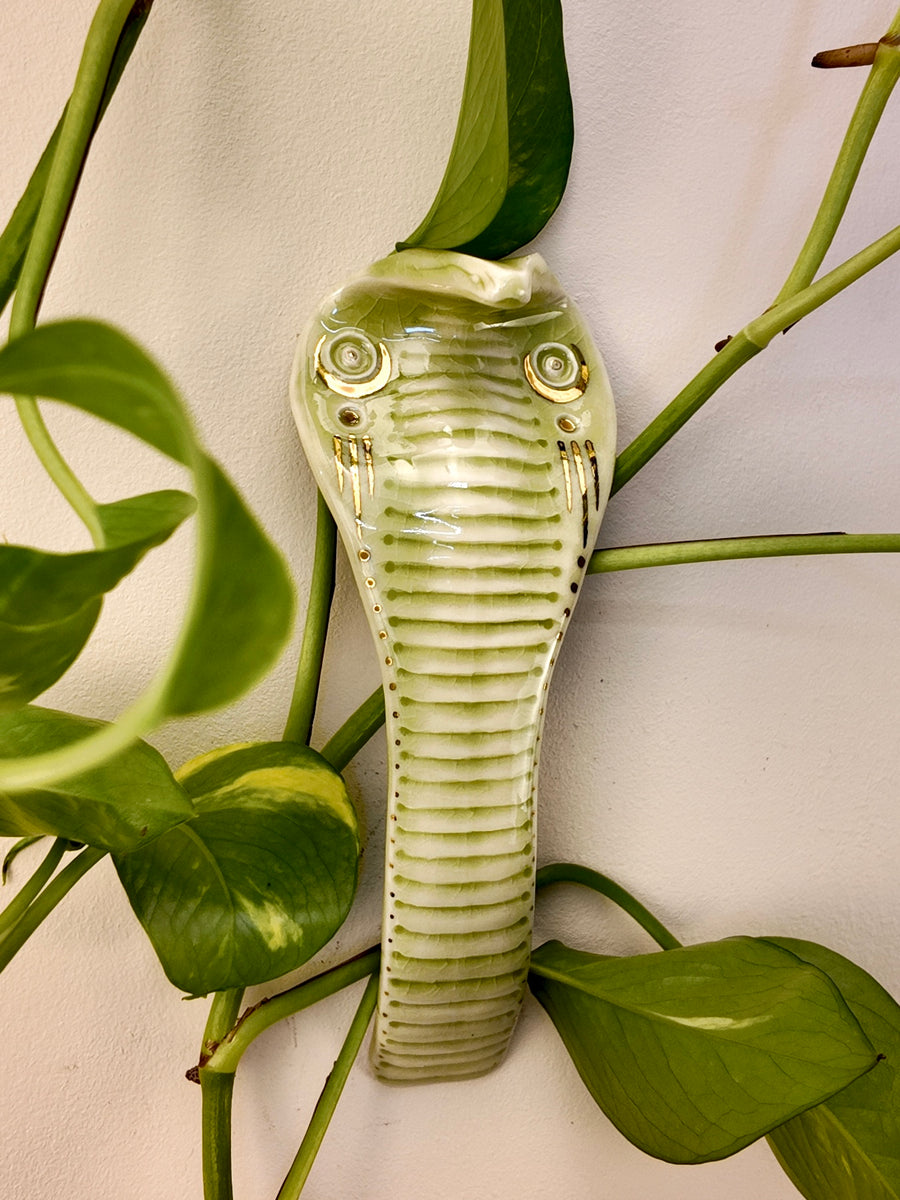 A ceramic wall-mounted snake figurine with a glossy finish, styled among green houseplant leaves against a plain off-white wall.