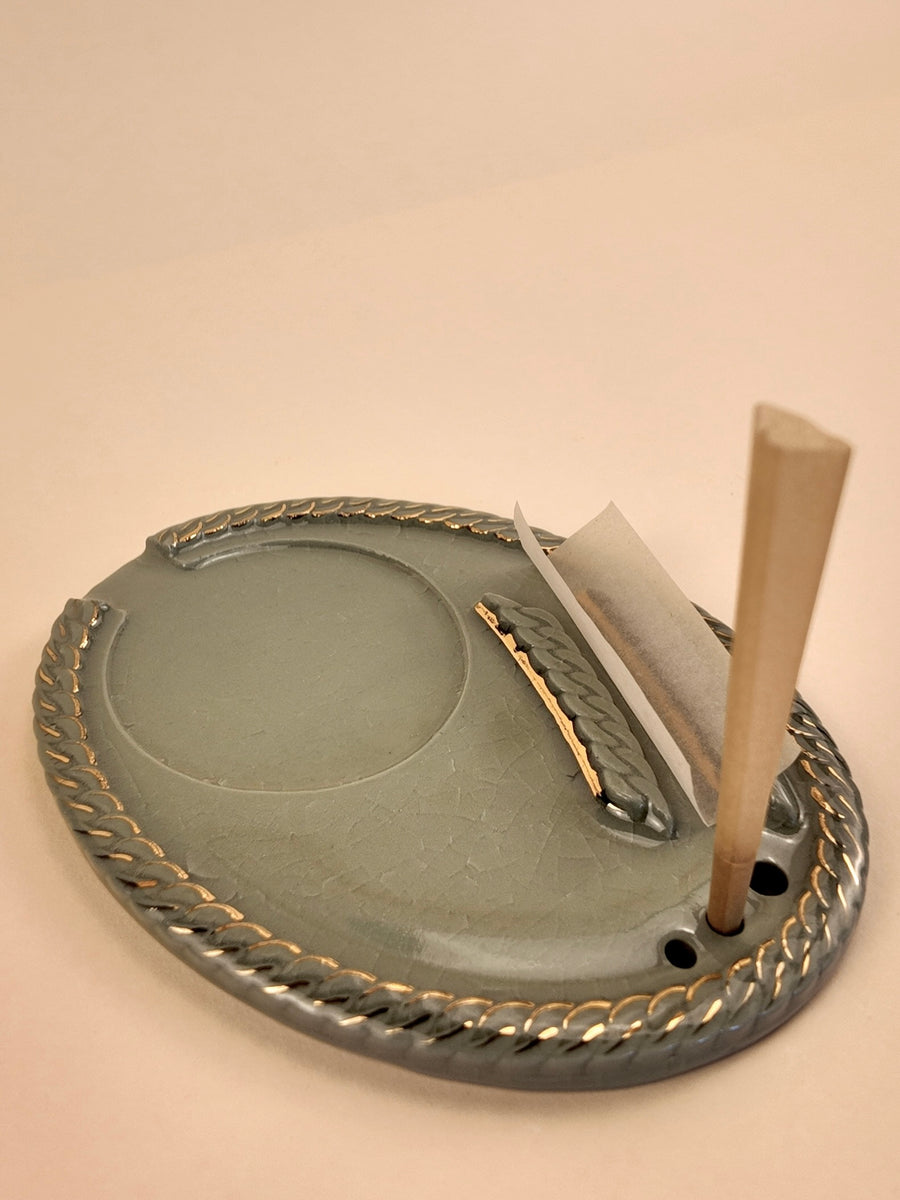 A round gray porcelain smoking tray with a braided gold rim, shown on a plain beige background. A rolling paper cone and a wooden rolling pin are placed inside the tray.