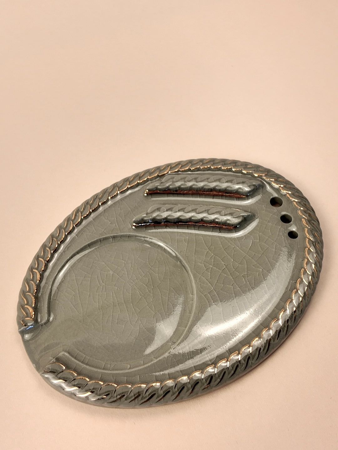 An oval porcelain ashtray with a textured metallic gray finish, featuring a braided rope border, two recessed slots for cigarettes, and three small drainage holes, set against a solid pale peach background.
