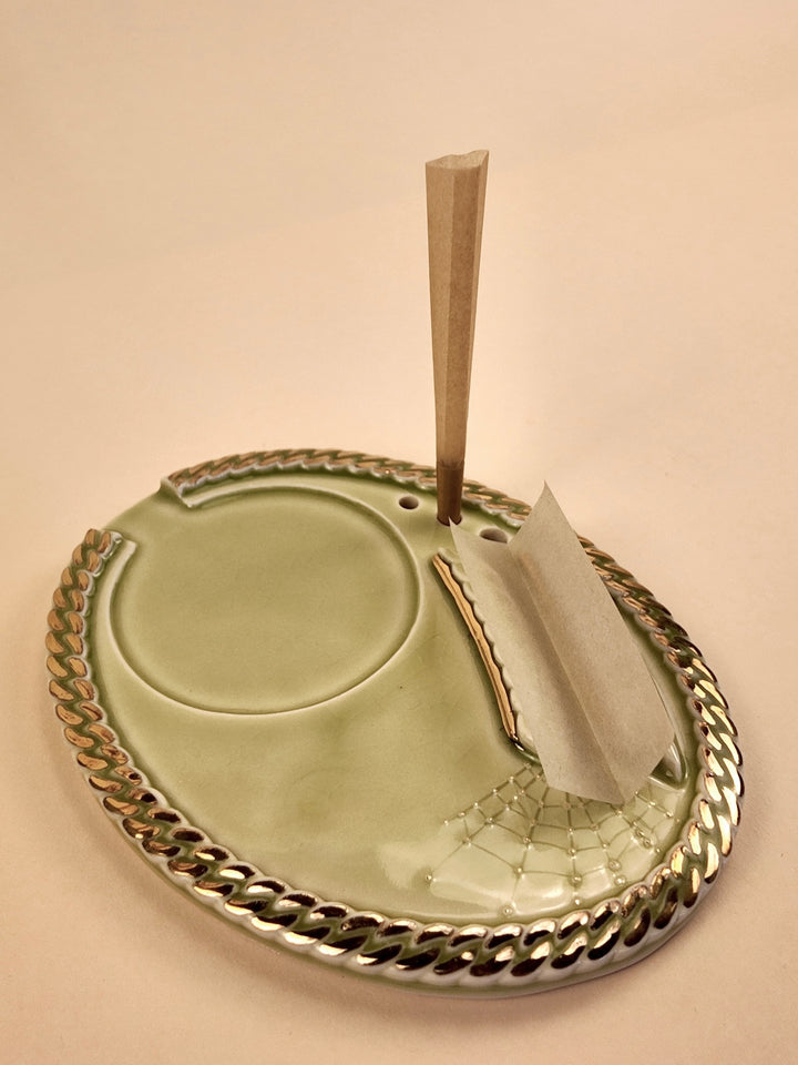 An oval porcelain ashtray with a braided metallic gold rim, featuring a circular recessed area, a paper slot with a rolled cigarette holder, and a vertical wooden cigarette stick standing upright, all on a warm beige background.