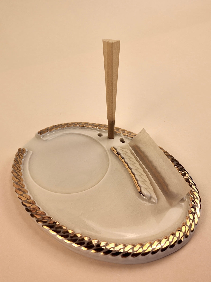 An oval porcelain ashtray with a braided metallic gold rim, featuring a circular recessed area, a paper slot with a rolled cigarette holder, and a vertical wooden cigarette stick standing upright, all on a warm beige background.