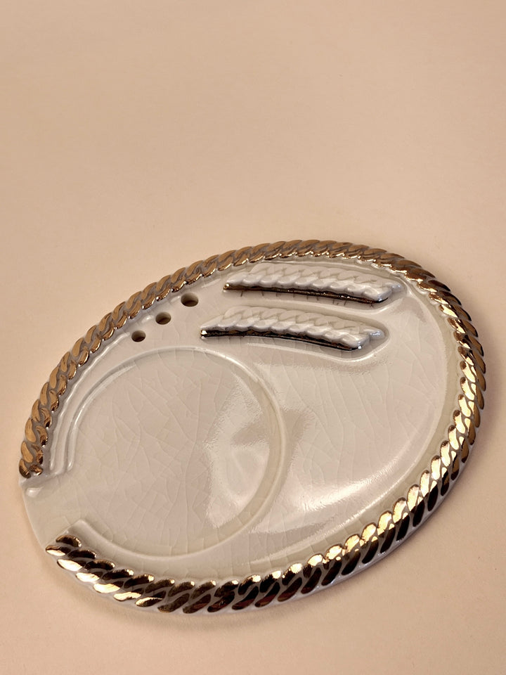 An oval porcelain ashtray with a braided metallic gold rim, featuring a circular recessed area, a paper slot, all on a warm beige background.