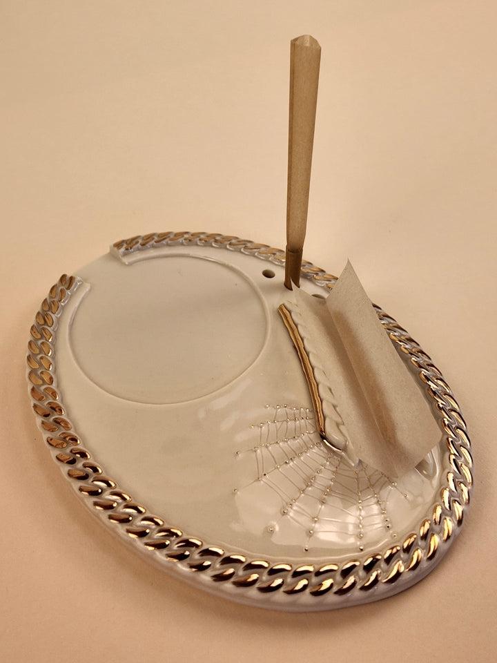 An oval white porcelain rolling tray with a metallic gold chain-link border, featuring a spiderweb design embossed in the center and a gold-toned cigarette paper holder with a wick, set against a solid beige background.
