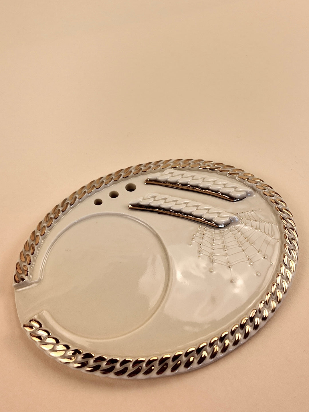 An oval white porcelain rolling tray with a metallic gold chain-link border, featuring a spiderweb design embossed in the center and a gold-toned cigarette papers holder, set against a solid beige background.