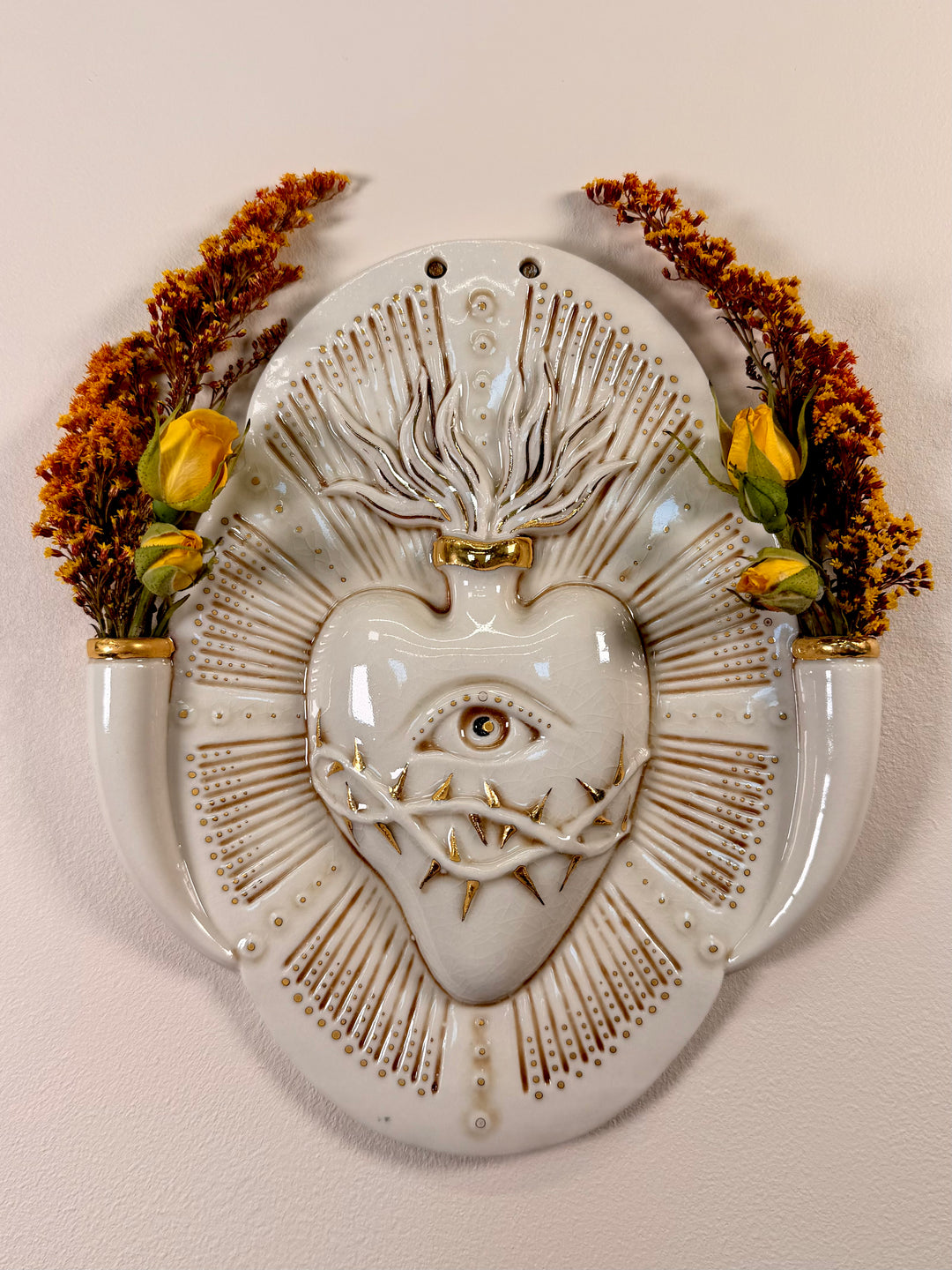 A decorative porcelain wall plaque or vase featuring a stylized heart with an eye and gold accents, styled with dried flowers in a matching ceramic vase against a neutral background.