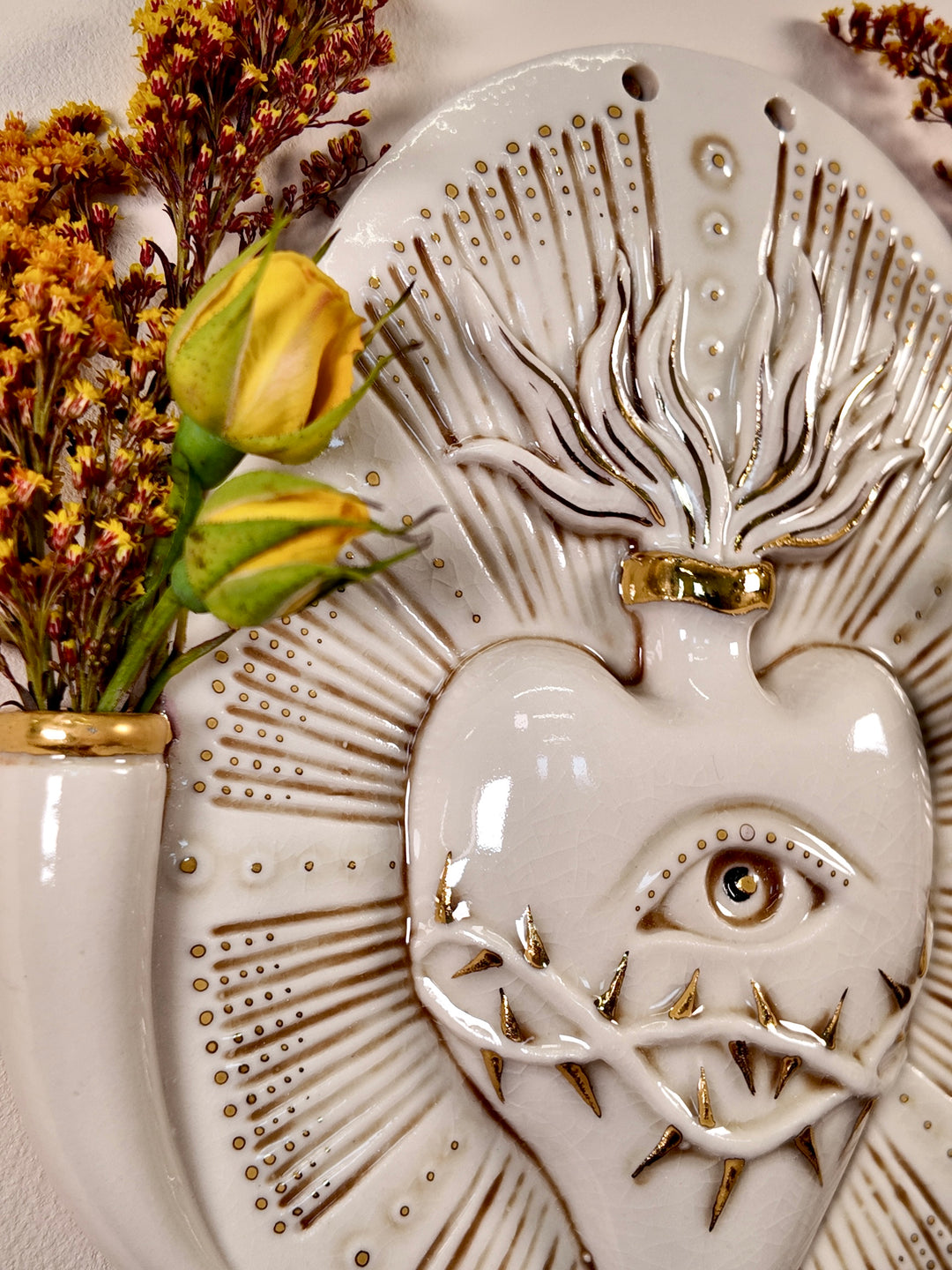 A decorative porcelain wall plaque or vase featuring a stylized heart with an eye and gold accents, styled with dried flowers in a matching ceramic vase against a neutral background.