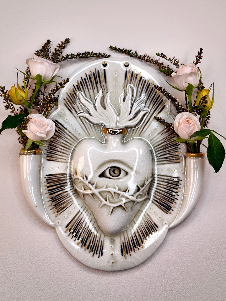 A white porcelain wall plaque or vase featuring a stylized heart with an eye, wrapped in thorny details, with gold accents and fluted handles on the sides holding pale pink roses and greenery, mounted on a light beige wall.