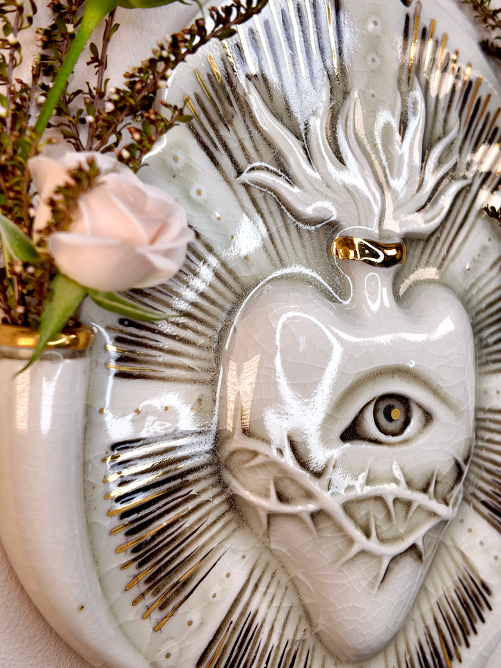 A close-up of a decorative porcelain wall piece featuring a stylized human eye with a thorny crown, surrounded by radiating textured rays and a pale pink rose in a white vase.