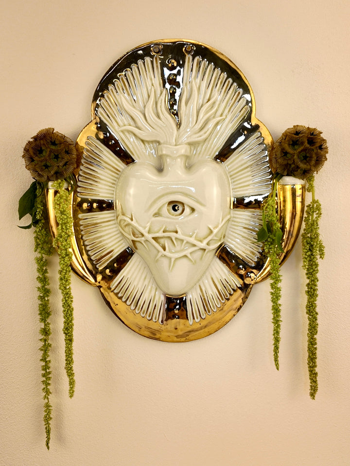 A decorative porcelain wall plaque featuring a stylized white heart with an eye and thorn details, surrounded by a gold-toned border, mounted on a beige wall with hanging green plants.