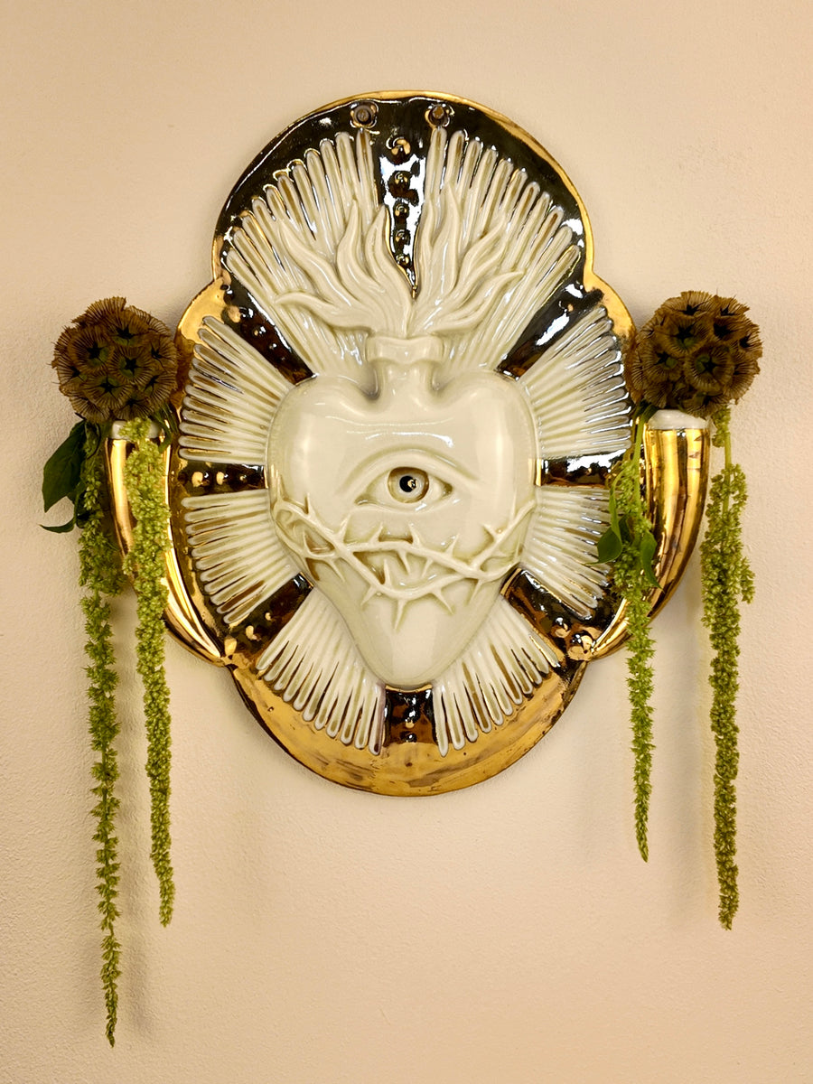 A decorative porcelain wall plaque featuring a stylized white heart with an eye and thorn details, surrounded by a gold-toned border, mounted on a beige wall with hanging green plants.