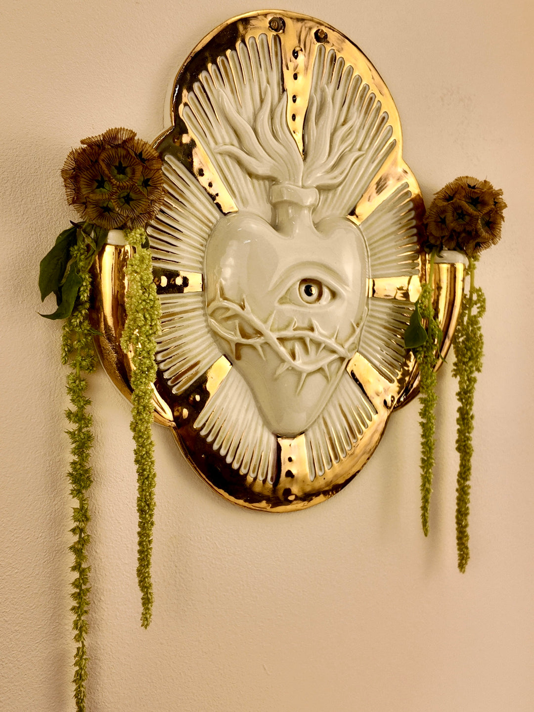 A decorative porcelain wall plaque featuring a stylized white heart with an eye and thorn details, surrounded by a gold-toned border, mounted on a beige wall with hanging green plants.