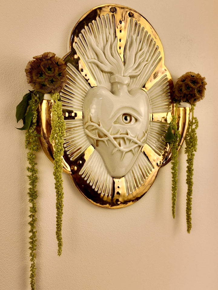 A decorative porcelain wall plaque featuring a stylized white heart with an eye and thorn details, surrounded by a gold-toned border, mounted on a beige wall with hanging green plants.