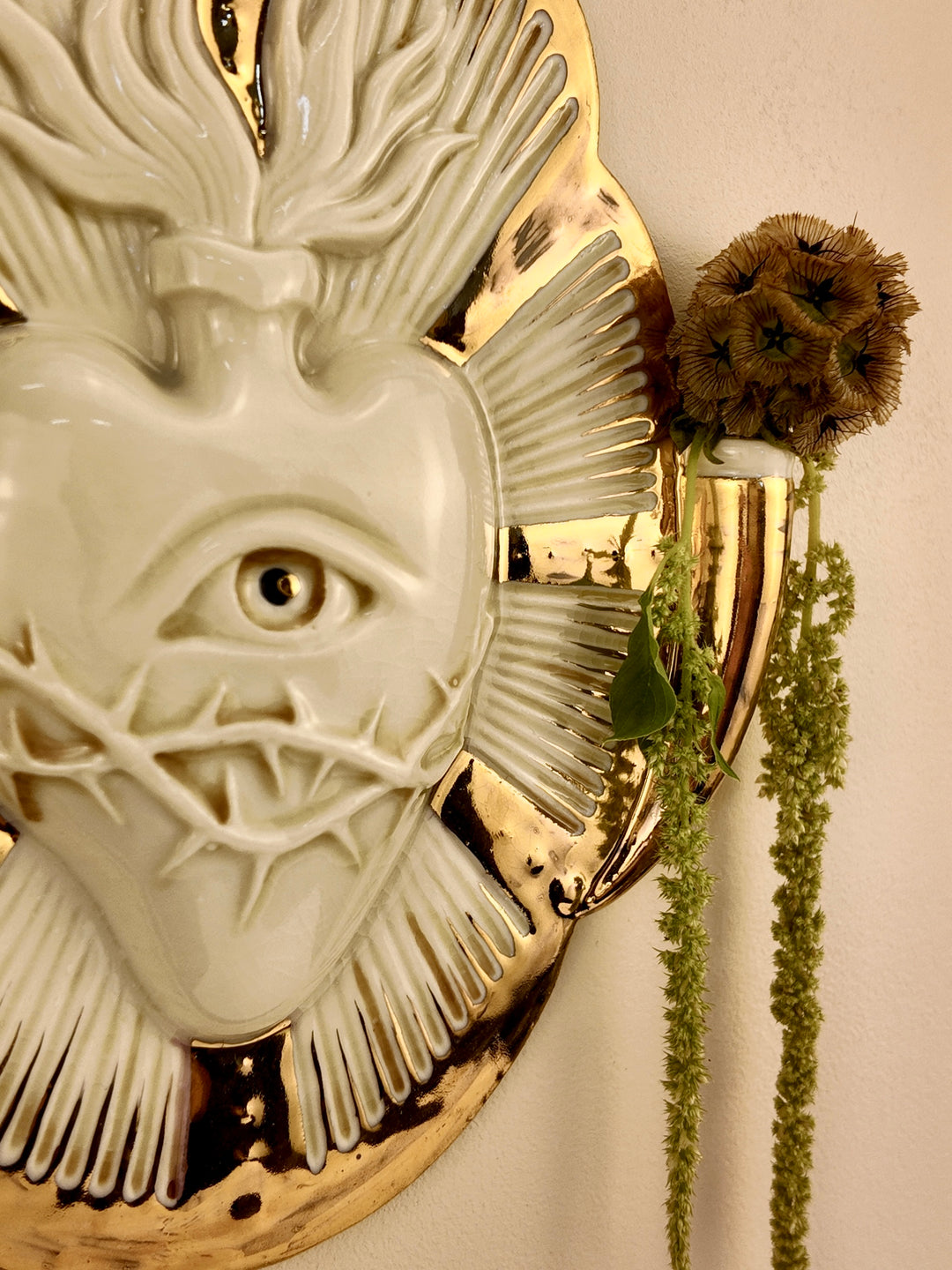 A close-up of a decorative wall-mounted porcelain vase featuring a white heart-like form with an eye and radiating textured details, set against a gold-toned circular base. A dried brown flower and green trailing stems are placed beside it against a plain light wall.