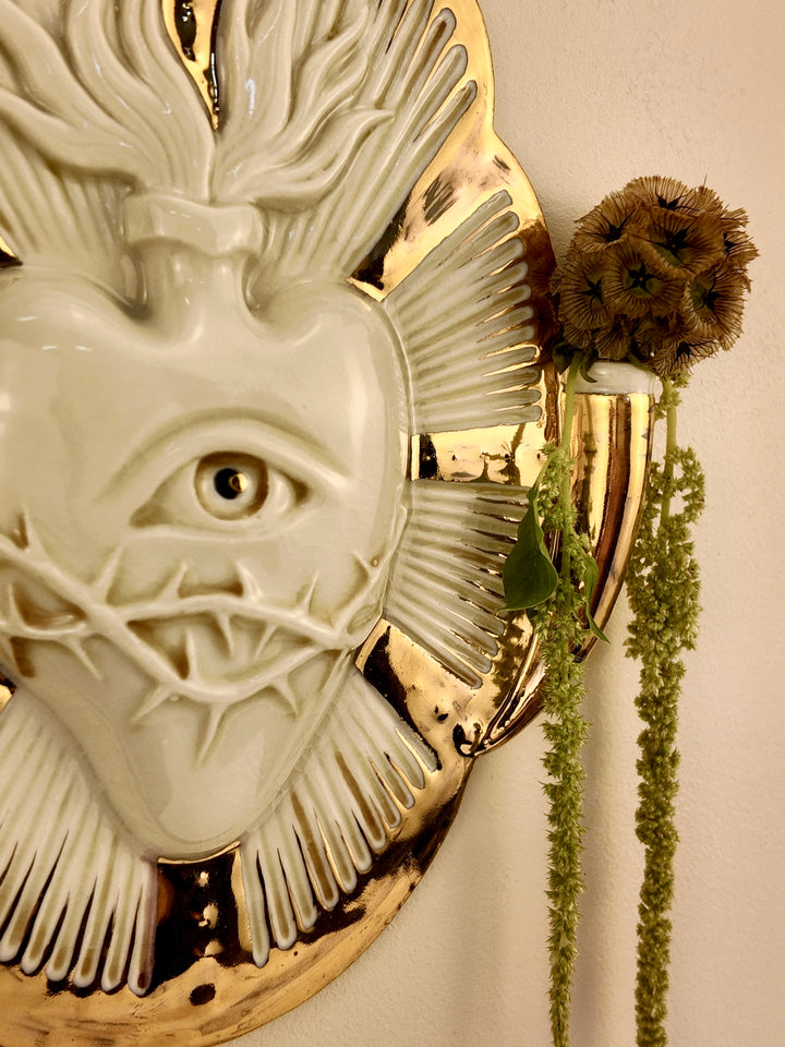 A close-up of a decorative wall-mounted porcelain vase featuring a white heart-like form with an eye and radiating textured details, set against a gold-toned circular base. A dried brown flower and green trailing stems are placed beside it against a plain light wall.