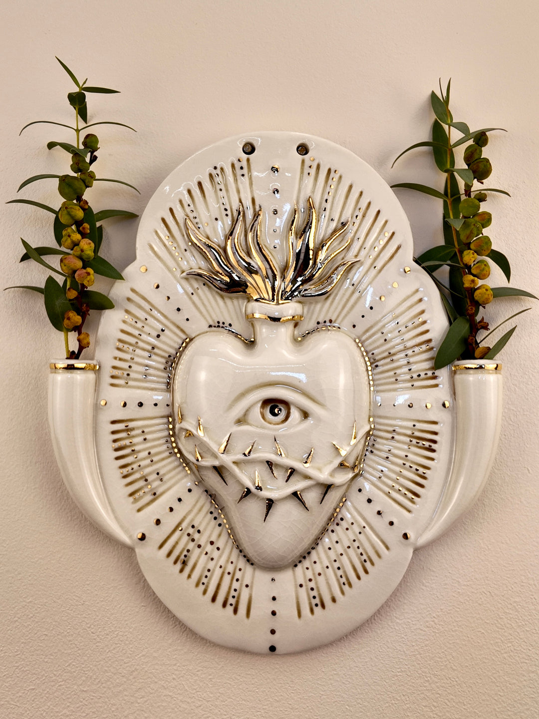 A decorative porcelain wall plaque featuring a stylized white heart with an eye and thorn details, surrounded by a gold-toned details, mounted on a beige wall with reference botanicals