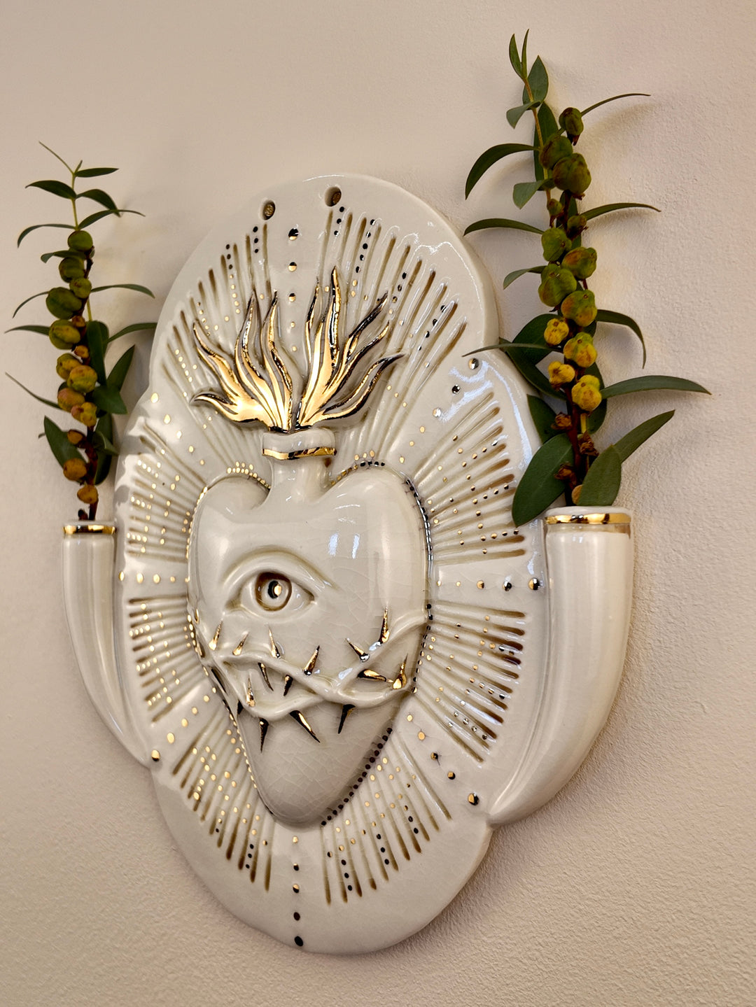A decorative porcelain wall plaque featuring a stylized white heart with an eye and thorn details, surrounded by a gold-toned details, mounted on a beige wall with reference botanicals