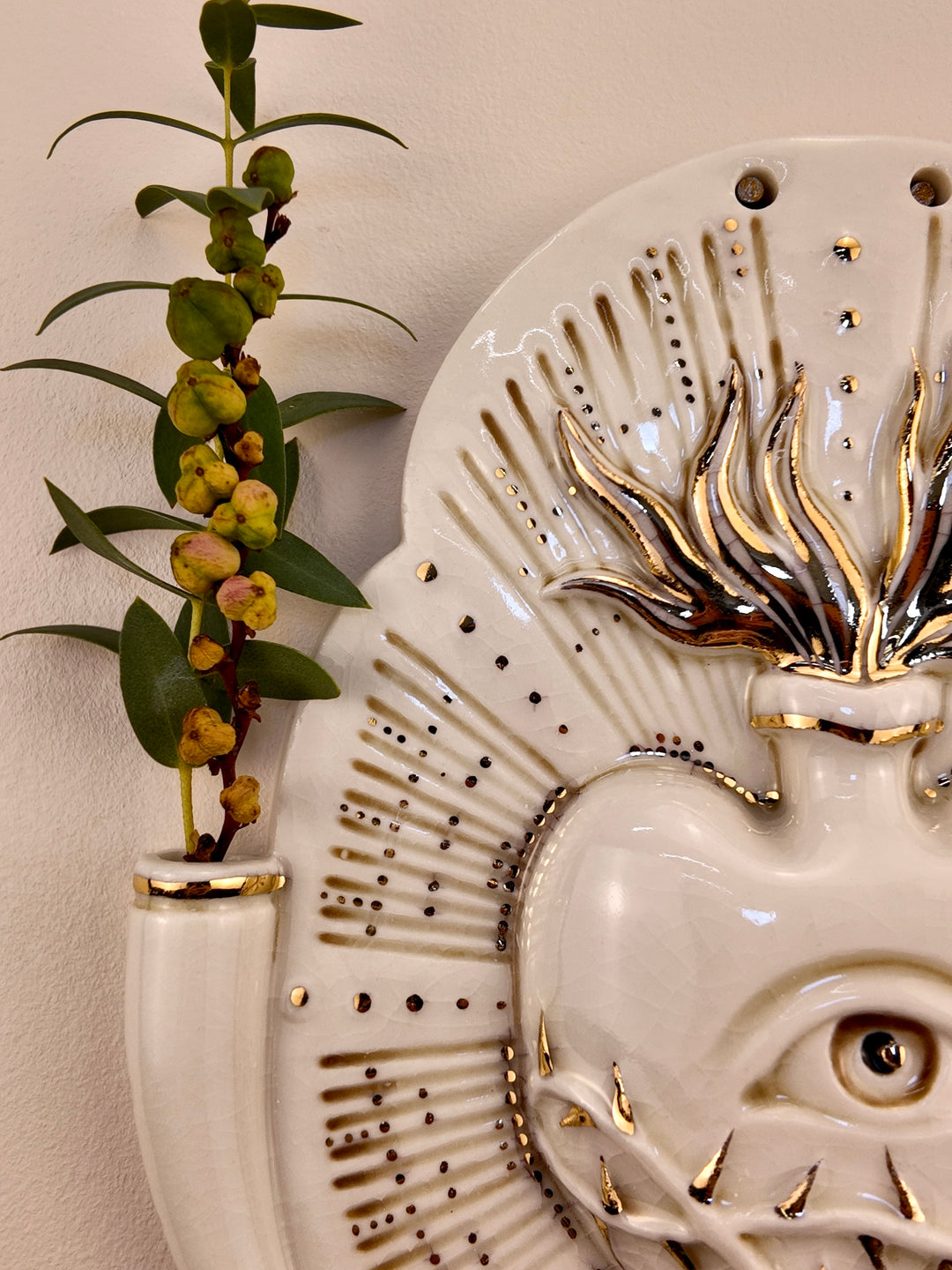 A close up of a decorative porcelain wall plaque featuring a stylized white heart with an eye and thorn details, surrounded by a gold-toned details, mounted on a beige wall with reference botanicals