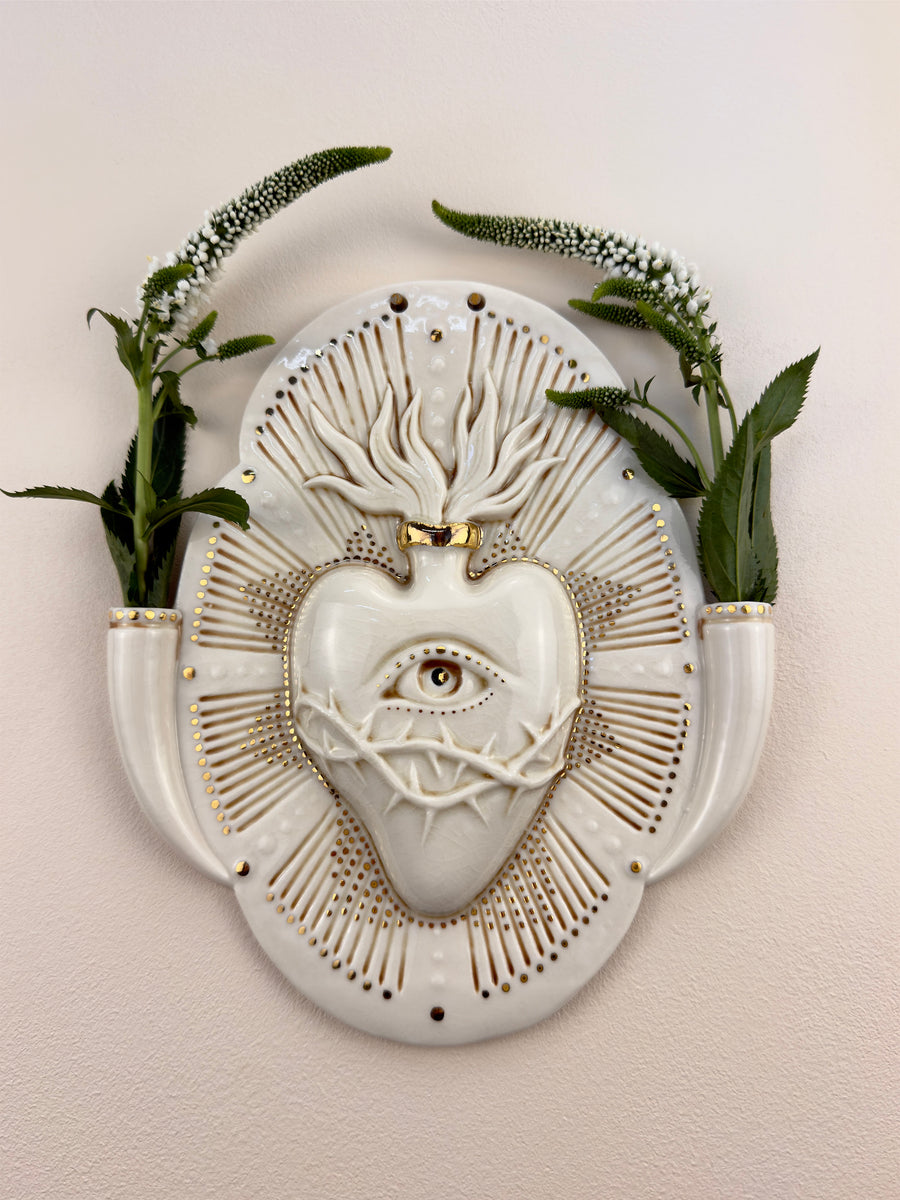 A decorative porcelain wall plaque featuring a stylized white heart with an eye and thorn details, surrounded by a gold-toned details, mounted on a beige wall with reference botanicals