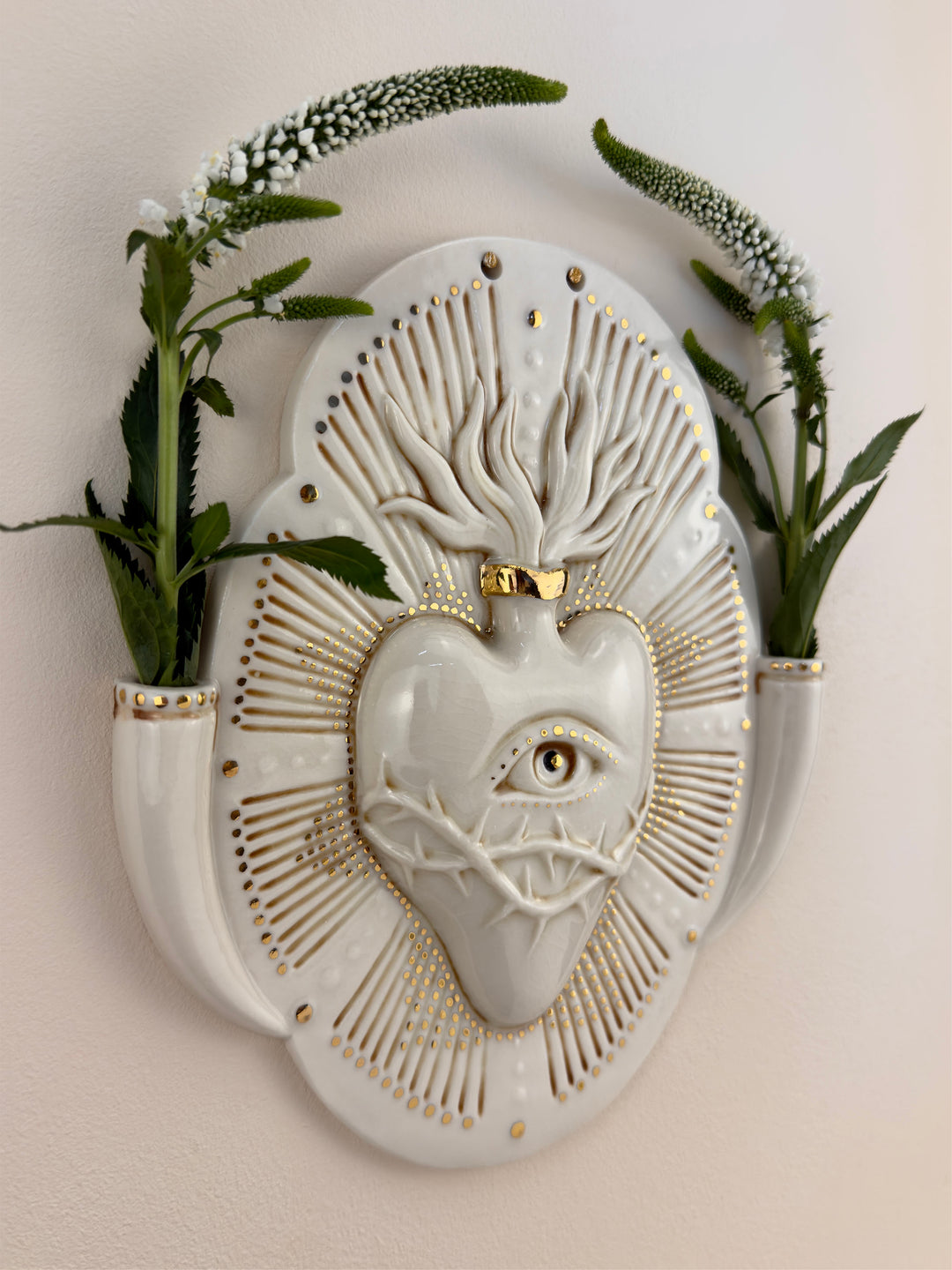 A decorative porcelain wall plaque featuring a stylized white heart with an eye and thorn details, surrounded by a gold-toned details, mounted on a beige wall with reference botanicals