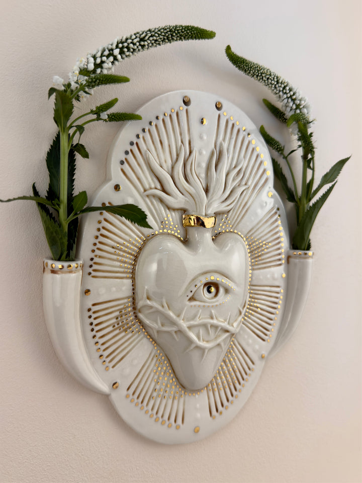 A decorative porcelain wall plaque featuring a stylized white heart with an eye and thorn details, surrounded by a gold-toned details, mounted on a beige wall with reference botanicals