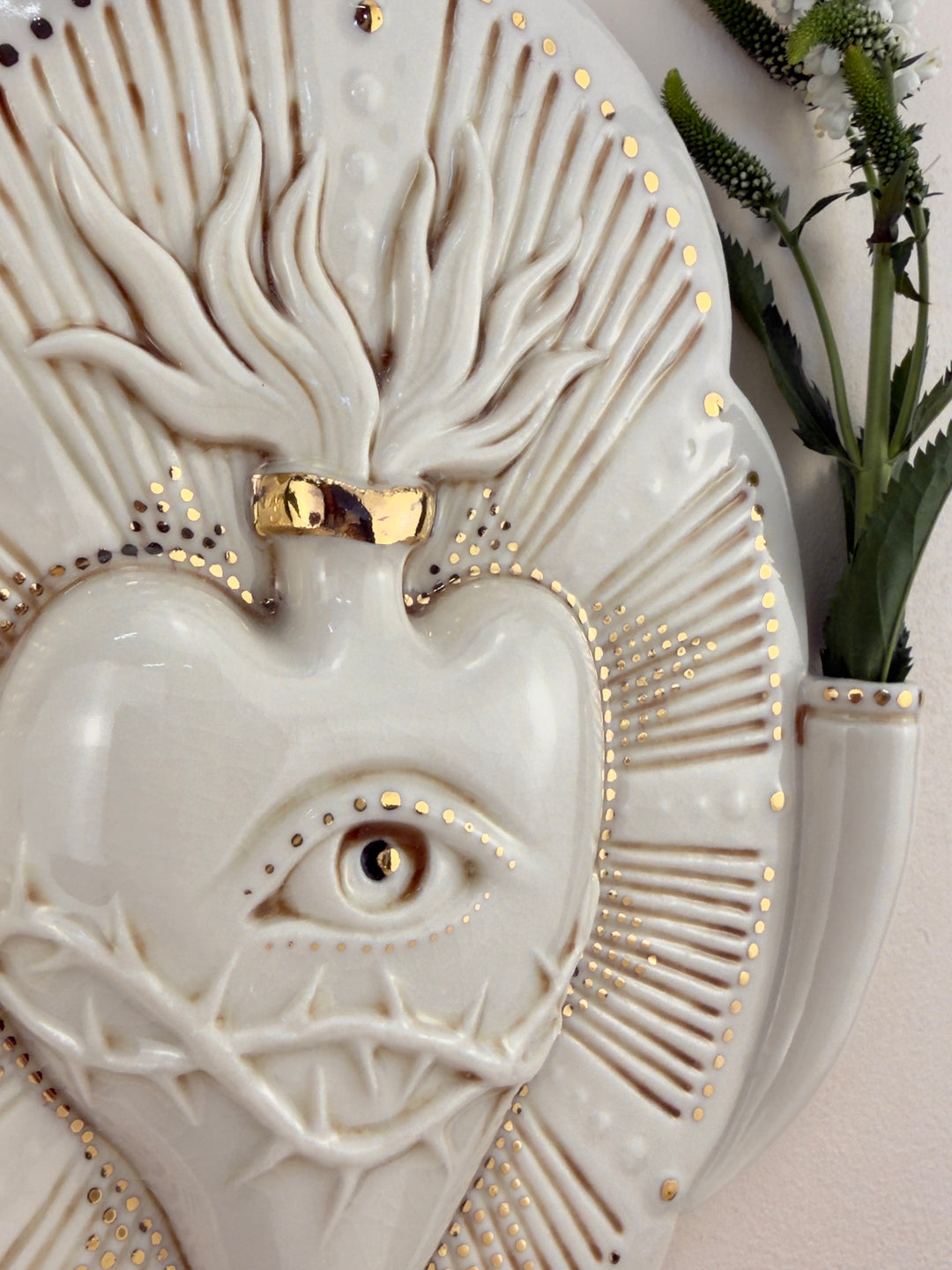 A close up of a decorative porcelain wall plaque featuring a stylized white heart with an eye and thorn details, surrounded by a gold-toned details, mounted on a beige wall with reference botanicals