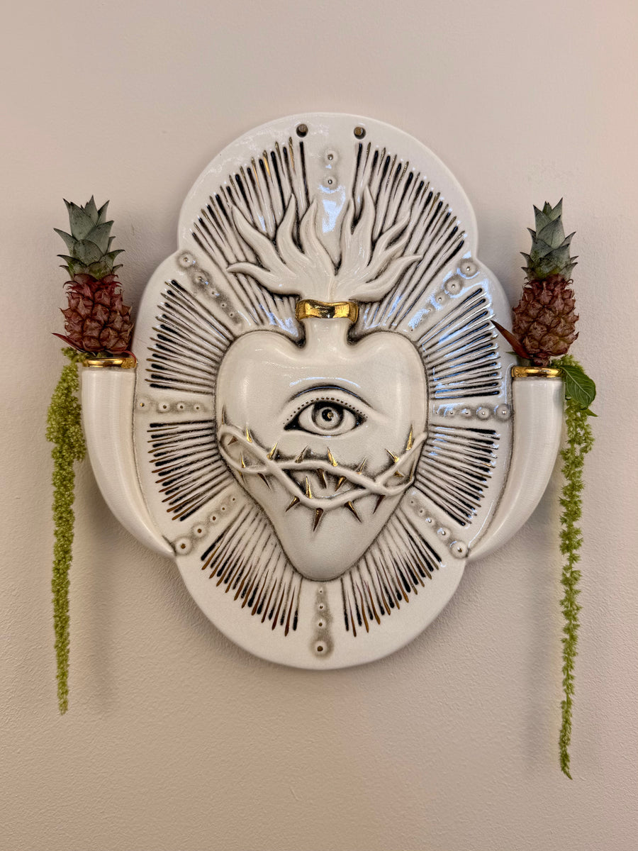 A decorative porcelain wall plaque featuring a stylized white heart with an eye and thorn details, mounted on a beige wall with reference botanicals