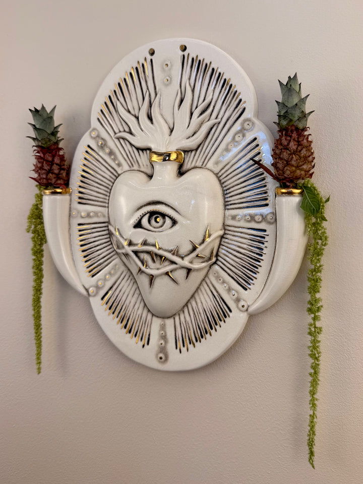 A decorative porcelain wall plaque featuring a stylized white heart with an eye and thorn details, mounted on a beige wall with reference botanicals