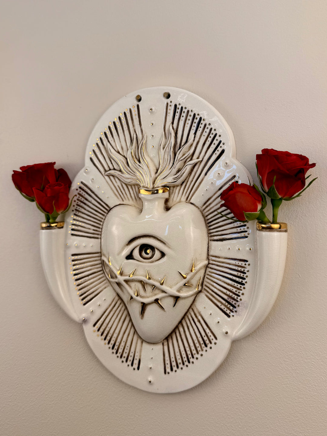 A decorative porcelain wall plaque featuring a stylized white heart with an eye and thorn details, mounted on a beige wall with reference Red Roses