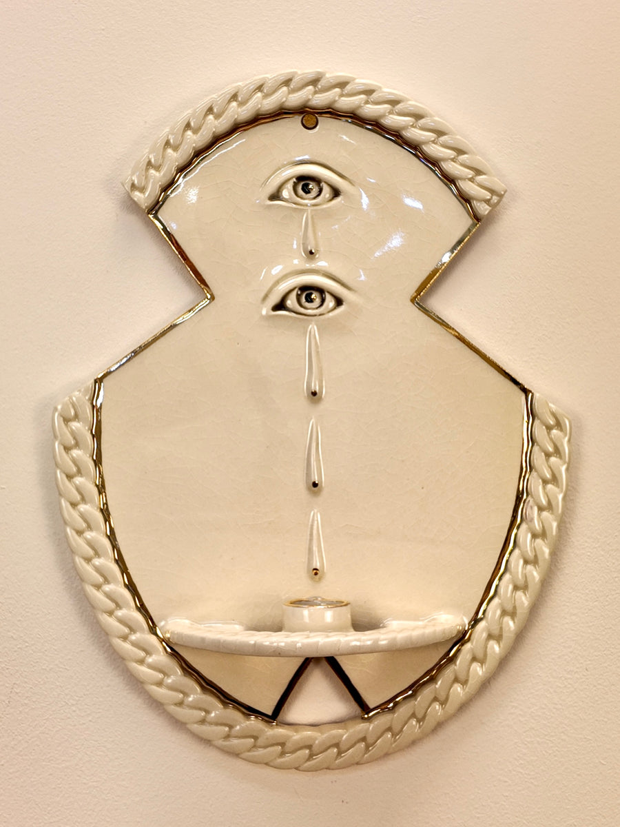 An off-white porcelain wall-mounted sculpture featuring two stylized human faces with eyes, a central vertical line of teardrop shapes, and braided rope-like borders around the top and bottom, shown against a plain beige wall.