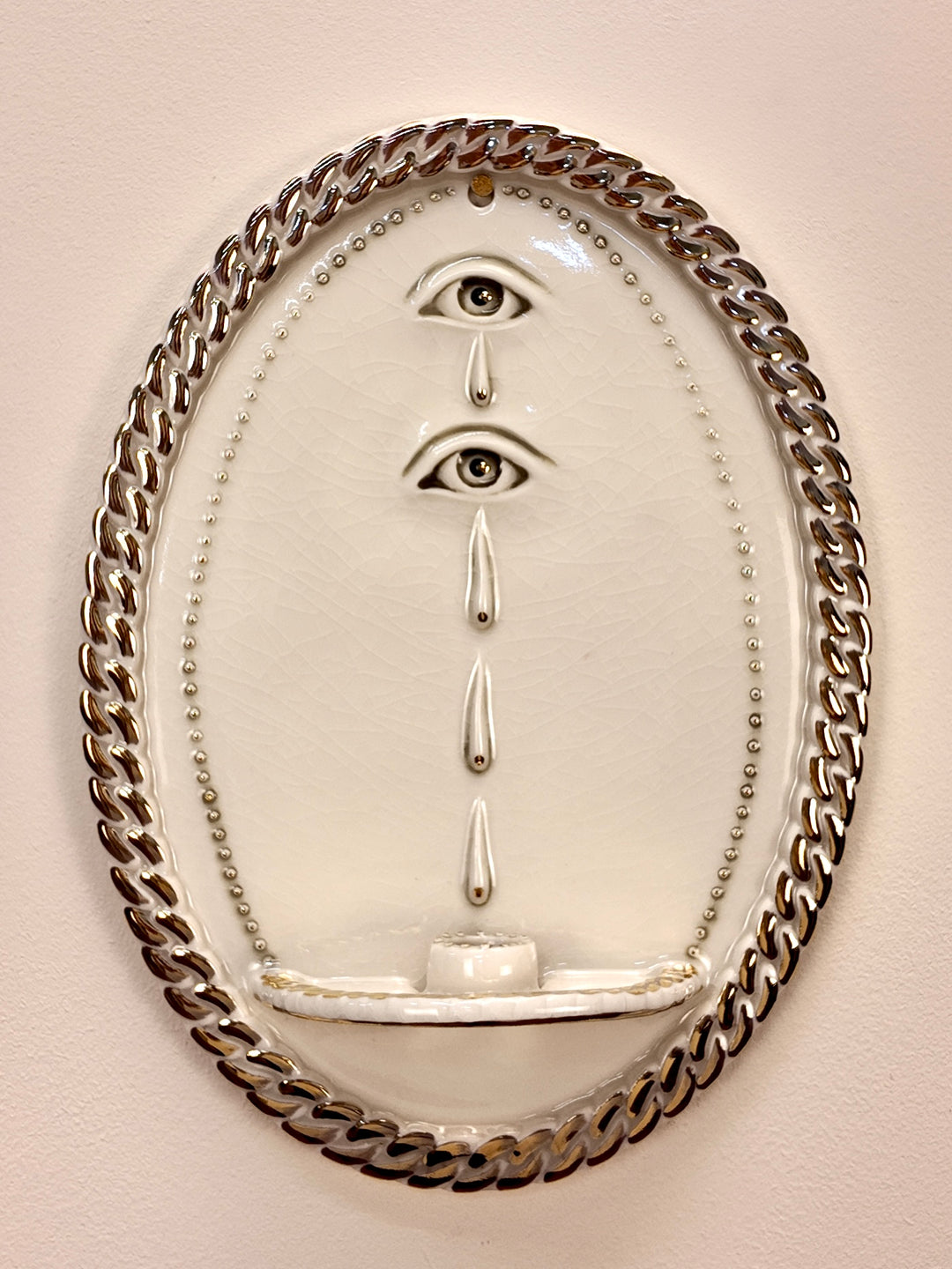 An oval wall-mounted porcelain sculpture featuring two stylized human eyes with dripping teardrop details, enclosed in a thick metallic chain border on a beige wall.