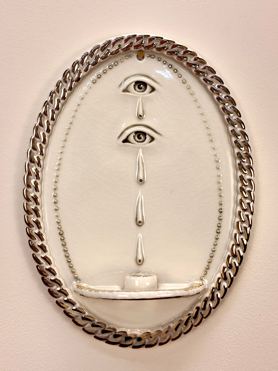 An oval wall-mounted porcelain sculpture featuring two stylized human eyes with dripping teardrop details, enclosed in a thick metallic chain border on a beige wall.
