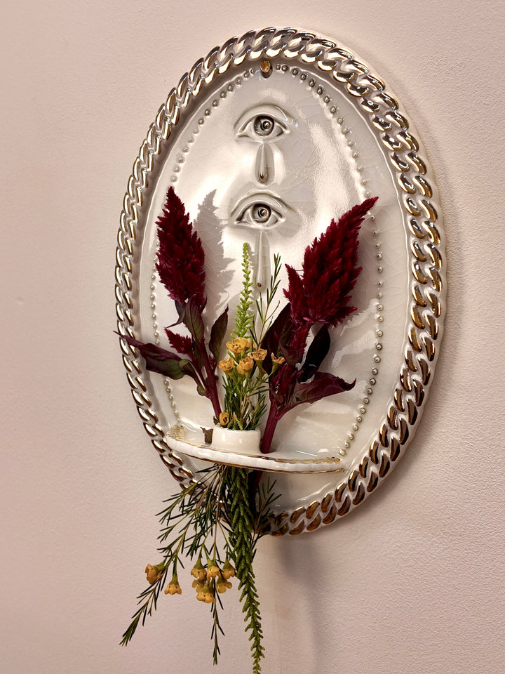 An oval wall-mounted porcelain sculpture featuring two stylized human eyes with dripping teardrop details, enclosed in a thick metallic chain border on a beige wall. Styled with dried flowers