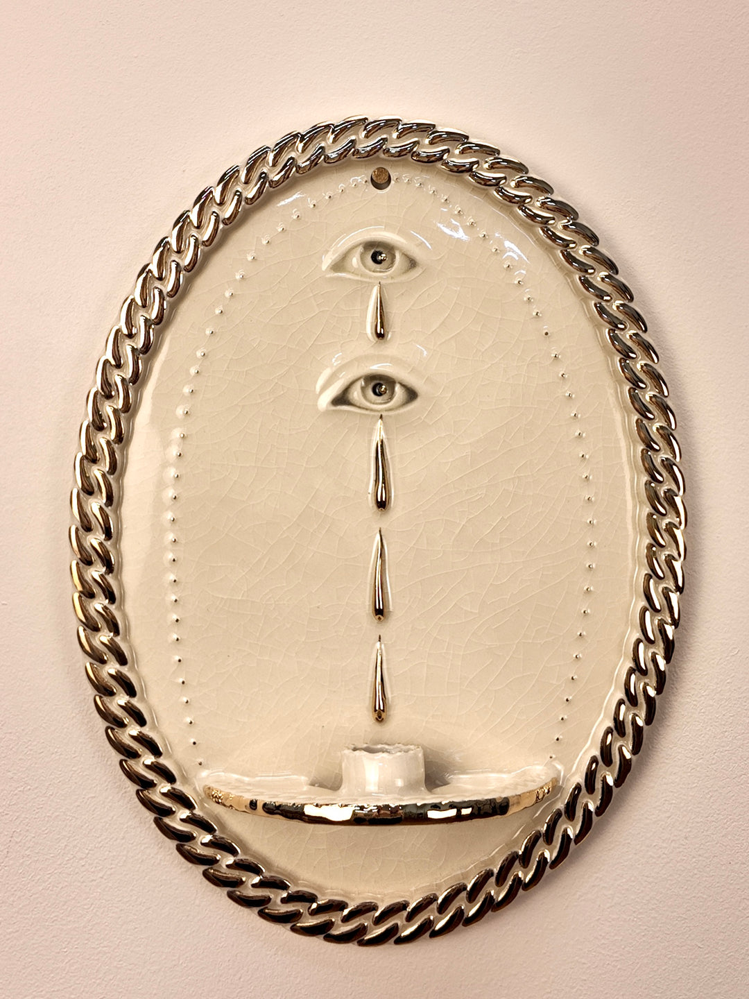 An oval wall-mounted stoneware and gold luster sculpture featuring two stylized eyes with dripping water motifs and a chain-link border, set against a plain light beige wall.