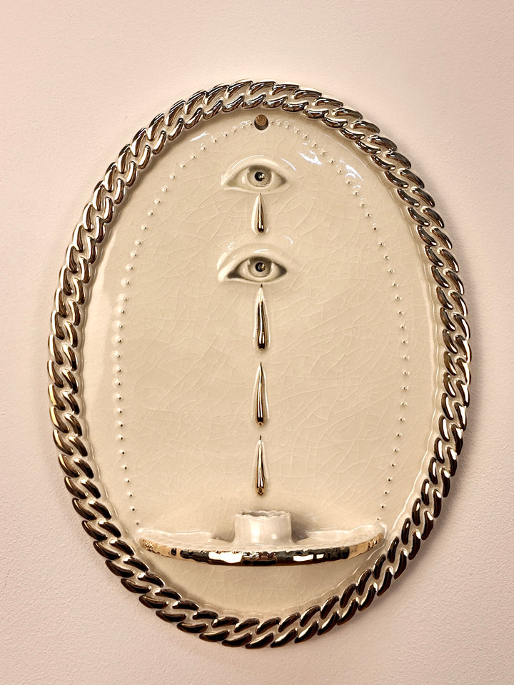 An oval wall-mounted stoneware and gold luster sculpture featuring two stylized eyes with dripping water motifs and a chain-link border, set against a plain light beige wall.