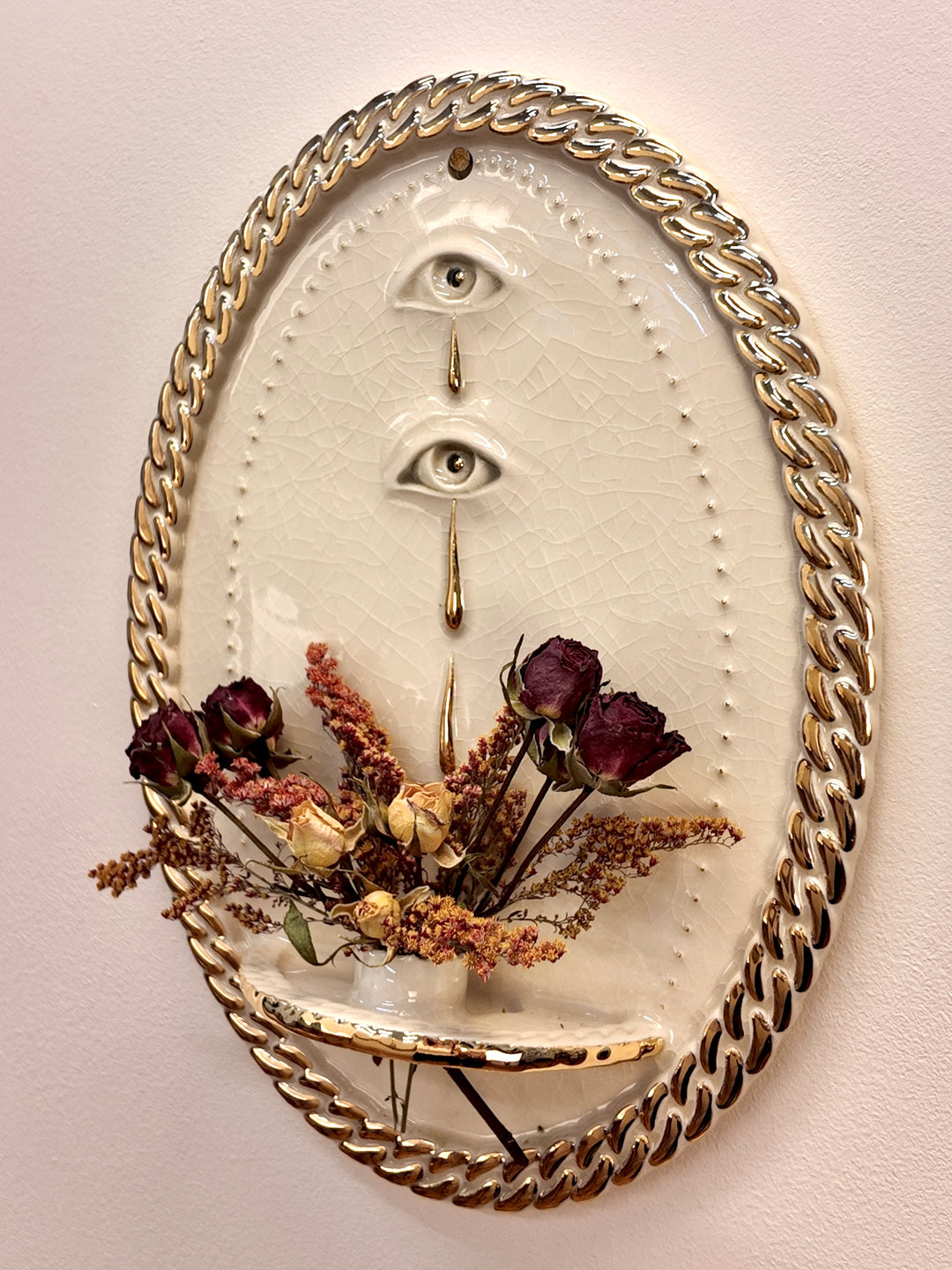 An oval stonware & gold luster wall art piece with a gold rope border, featuring two vertical eyes with tears and a small shelf holding dried burgundy roses and tan botanicals, mounted on a light beige wall.