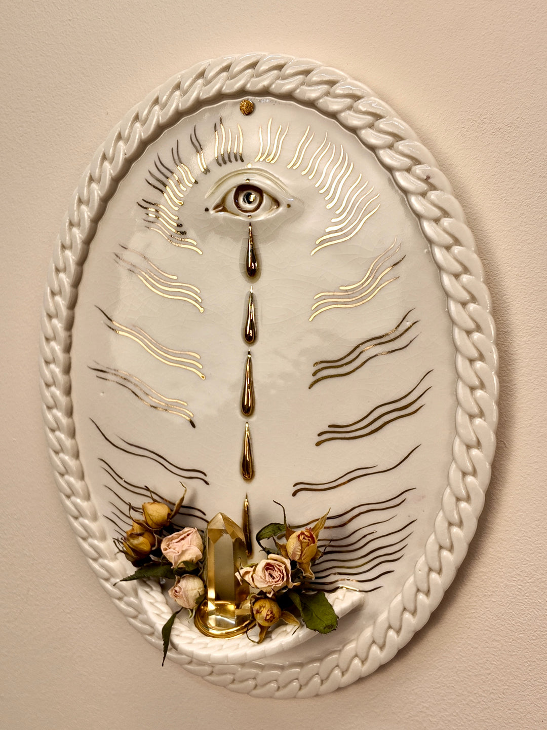 An oval porcelain & gold luster wall art piece featuring a white background with a braided rope border, a central eye motif, gold and black line details, dried roses, crystals, and a small gold candle holder at the bottom, mounted on a beige wall.