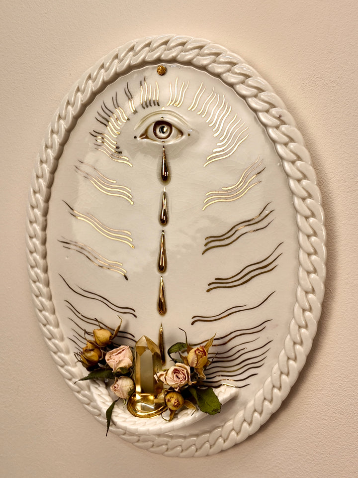 An oval porcelain & gold luster wall art piece featuring a white background with a braided rope border, a central eye motif, gold and black line details, dried roses, crystals, and a small gold candle holder at the bottom, mounted on a beige wall.