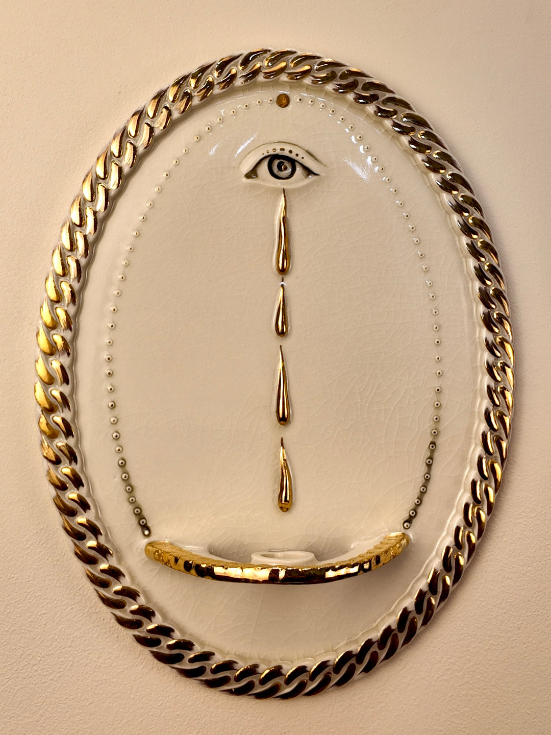 An oval wall-mounted porcelain and gold luster sculpture featuring two stylized eyes with dripping water motifs and a chain-link border, set against a plain light beige wall.