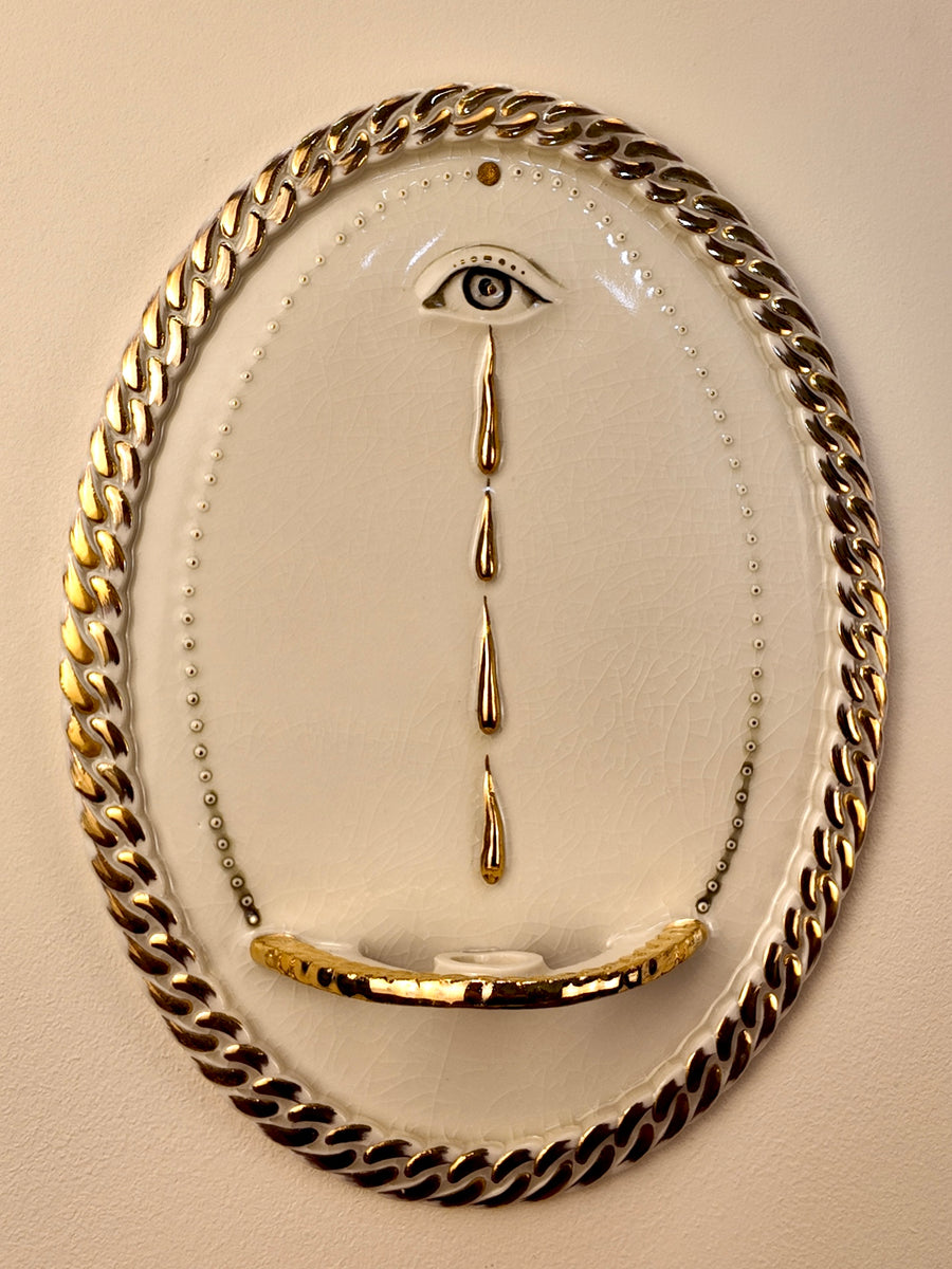 An oval wall-mounted porcelain and gold luster sculpture featuring two stylized eyes with dripping water motifs and a chain-link border, set against a plain light beige wall.