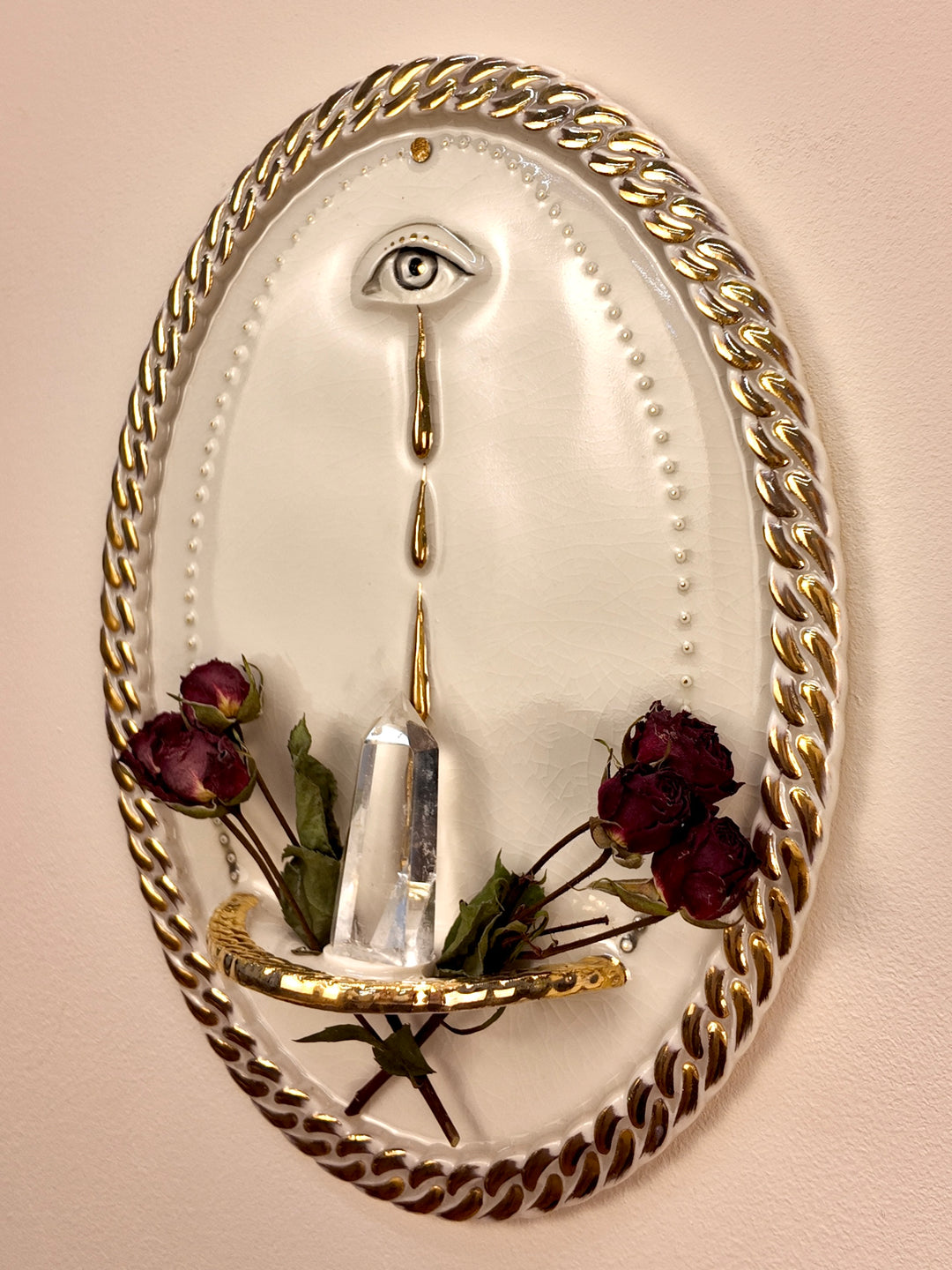 An oval wall-mounted porcelain and gold luster sculpture featuring two stylized eyes with dripping water motifs and a chain-link border, set against a plain light beige wall styled with dried roses