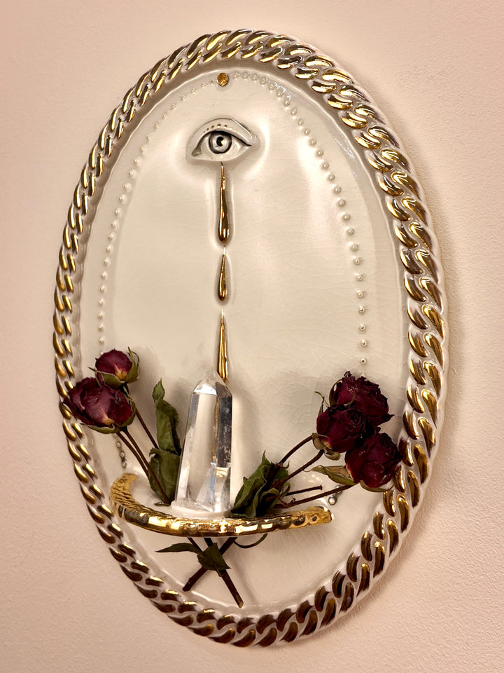 An oval wall-mounted porcelain and gold luster sculpture featuring two stylized eyes with dripping water motifs and a chain-link border, set against a plain light beige wall styled with dried roses