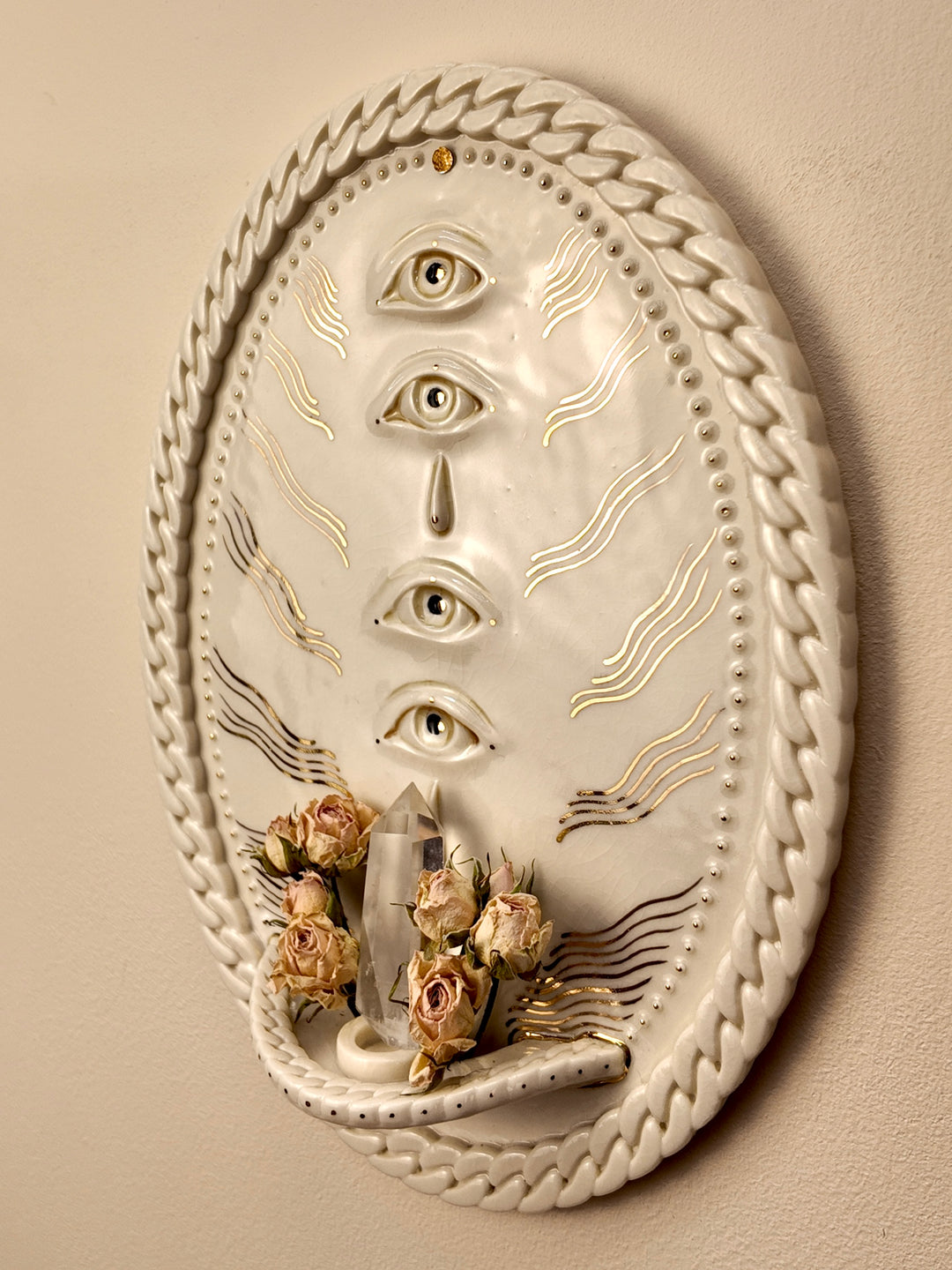 An oval white porcelain & gold luster wall-mounted sculpture featuring four stacked eyes, gold line details, a crystal cluster, and dried flowers at the base, set against a beige wall.